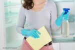 afvoergaatje koelkast verstopt: persoon voor koelkast met schoonmaakdoekje en sprayfles in handen