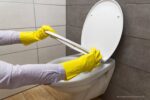 Gele aanslag op wc-bril verwijderen: een persoon met gele huishoudhandschoenen die een toiletbril omhoog houdt