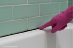 Schimmel op kitranden langs badrand in de badkamer, zwarte aanslag op de kit terwijl een hand met roze schoonmaakhandschoen de aangetaste rand aanwijst.