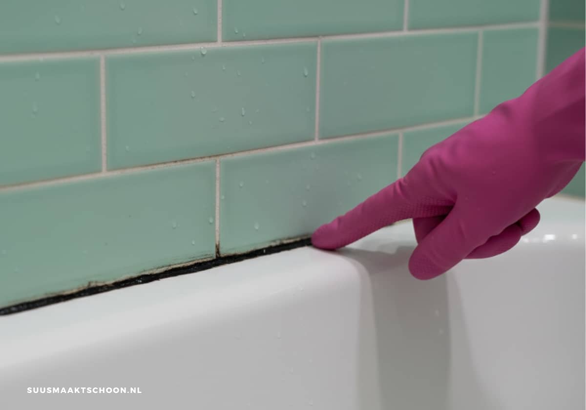 Schimmel op kitranden langs badrand in de badkamer, zwarte aanslag op de kit terwijl een hand met roze schoonmaakhandschoen de aangetaste rand aanwijst.